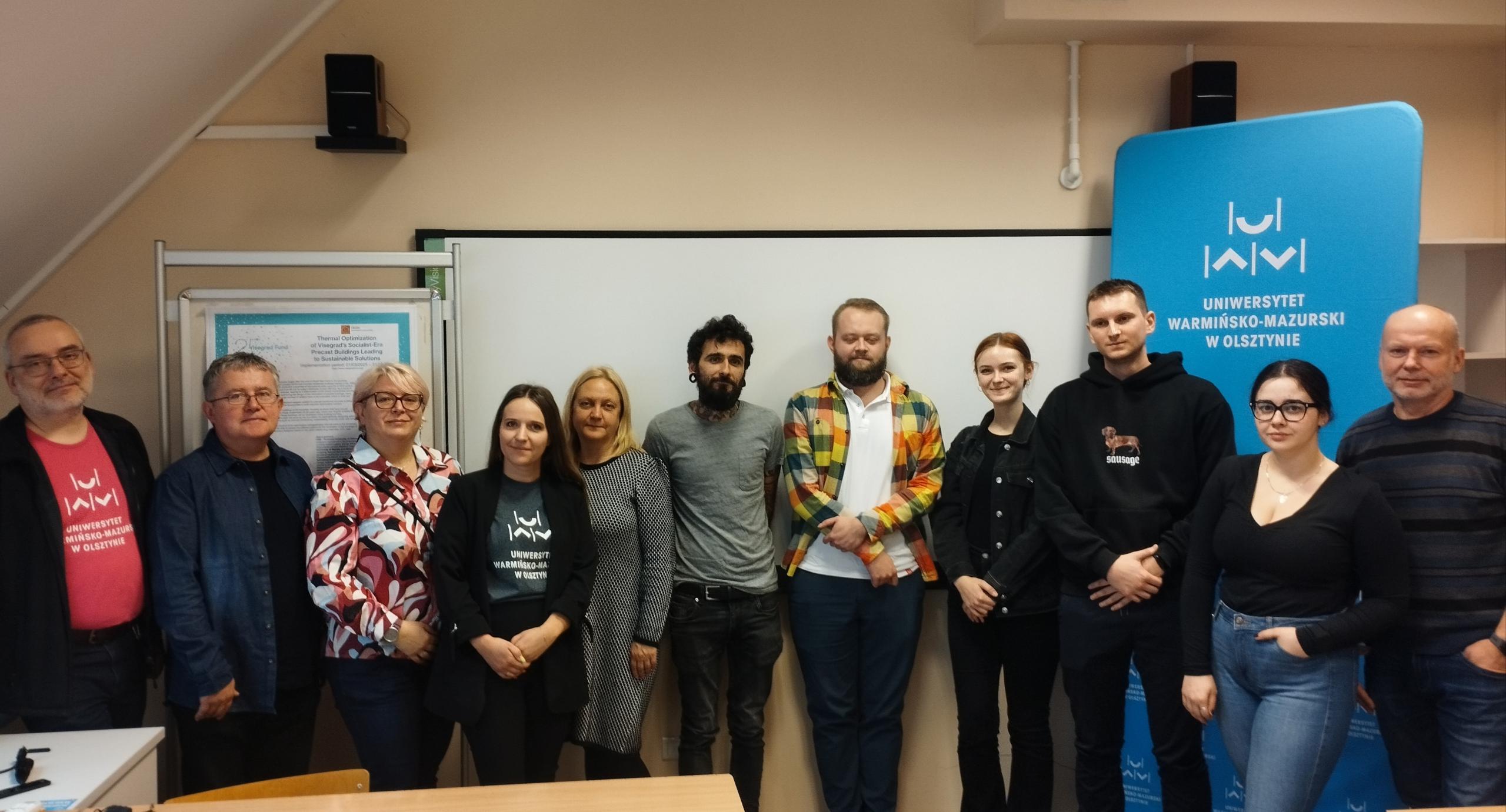 zdjęcie grupowe uczestników seminarium. Kobiety i mężczysni stoją w sali a w tle logotyp UWM