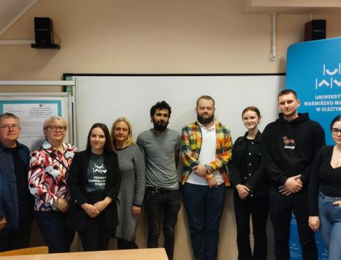 zdjęcie grupowe uczestników seminarium. Kobiety i mężczysni stoją w sali a w tle logotyp UWM